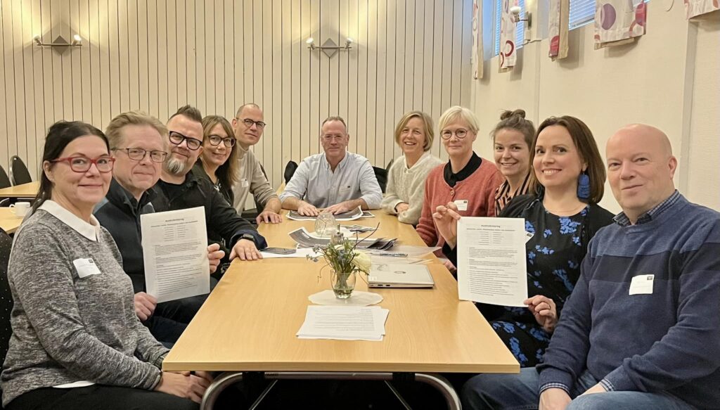 En gruppbild på de medverkade kultur- och musikskolorna i Västerbotten. De medverkande på bilden sitter placerade runt ett rektangulärt bord och alla ler. Två personer håller upp varsitt pappersdokumet. på bordet står en liten vas med blommor i.