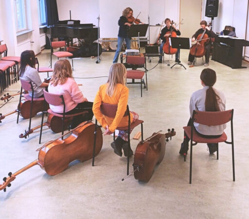 Barn provar på att spela cello. Fyra barn sitter med varsin cello bredvid sig på golvet, och tittar på en lärare som spelar fiol och två andra lärare spelar cello.
