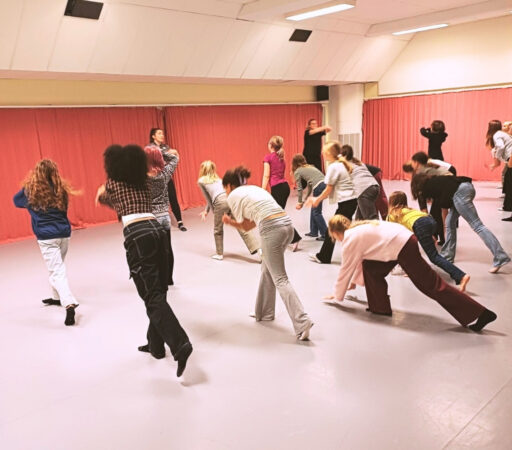 Barn provar på dans och står vända mot två lärare. Det är i ett stort upplyst rum med vitt tak och röda tygstycken på väggarna.