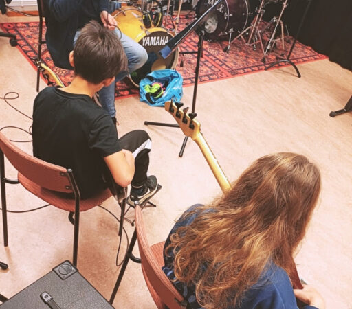Två barn sitter med ryggen åt och spelar bas. I bakgrunden syns delar av ett trumset.