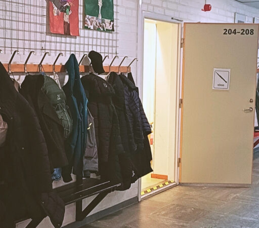 En korridor där krokar på vägen är fulla med ytterkläder. En sörj står öppen och på dörren sitter en lapp som det står "oboe" och visar även en bild på en oboe.